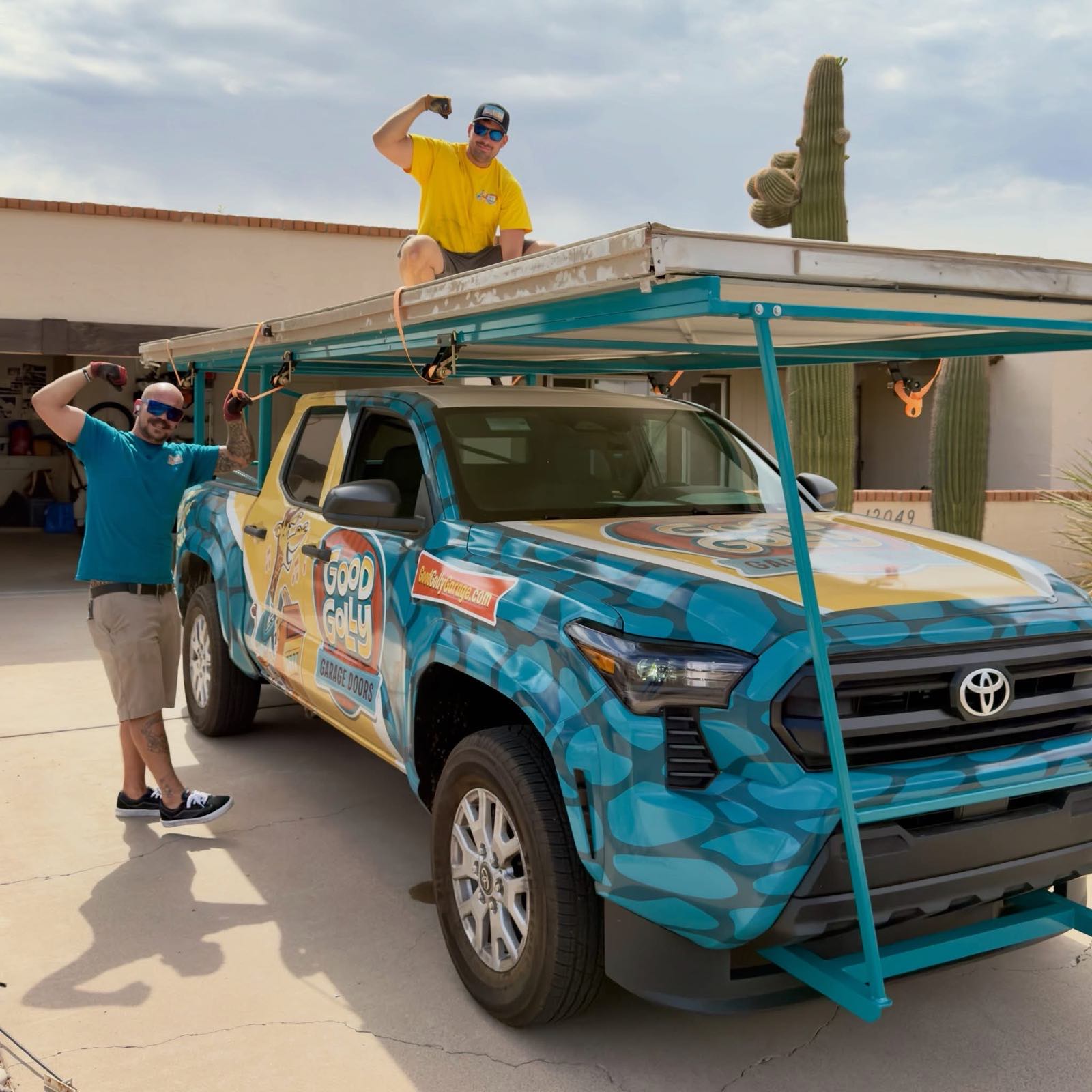 Two Good Golly technicians strapping removed garage door sections onto the rack of the branded truck in a Phoenix driveway, with a saguaro standing behind the house