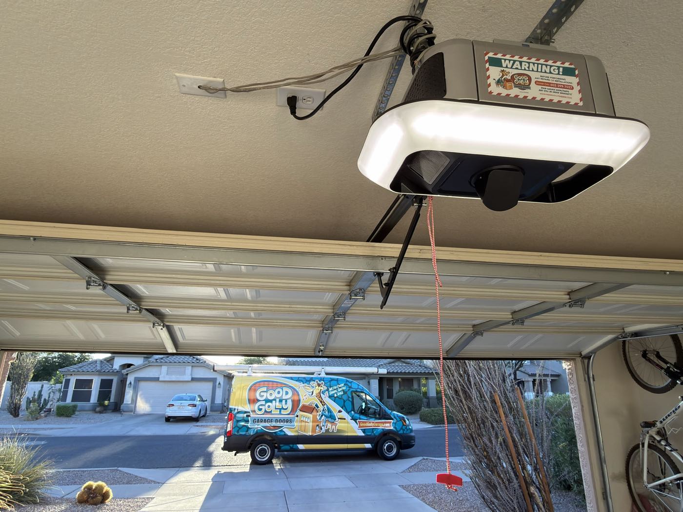 A serviced garage door opener carrying the Good Golly safety label in a Phoenix garage, with the branded van on the driveway outside
