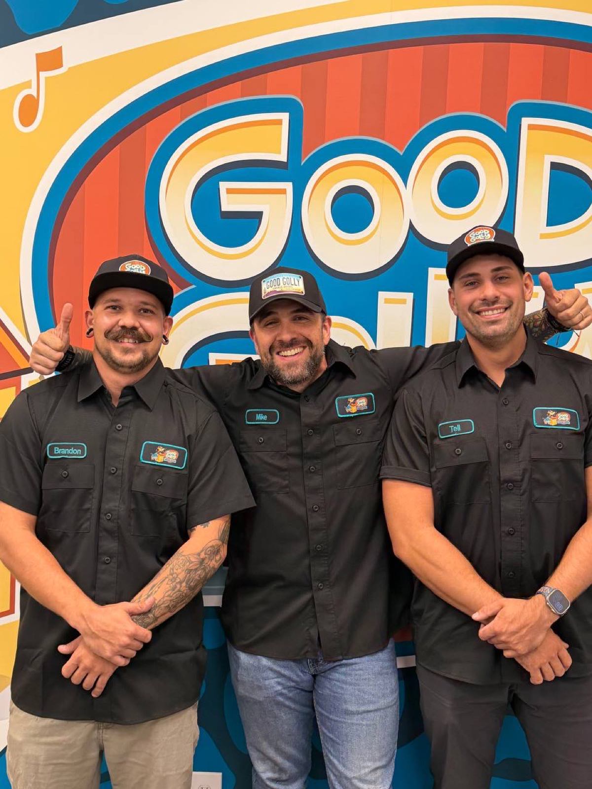 Three Good Golly Garage Doors team members in branded shirts and caps giving a thumbs up in front of the painted Good Golly mural at the Phoenix shop