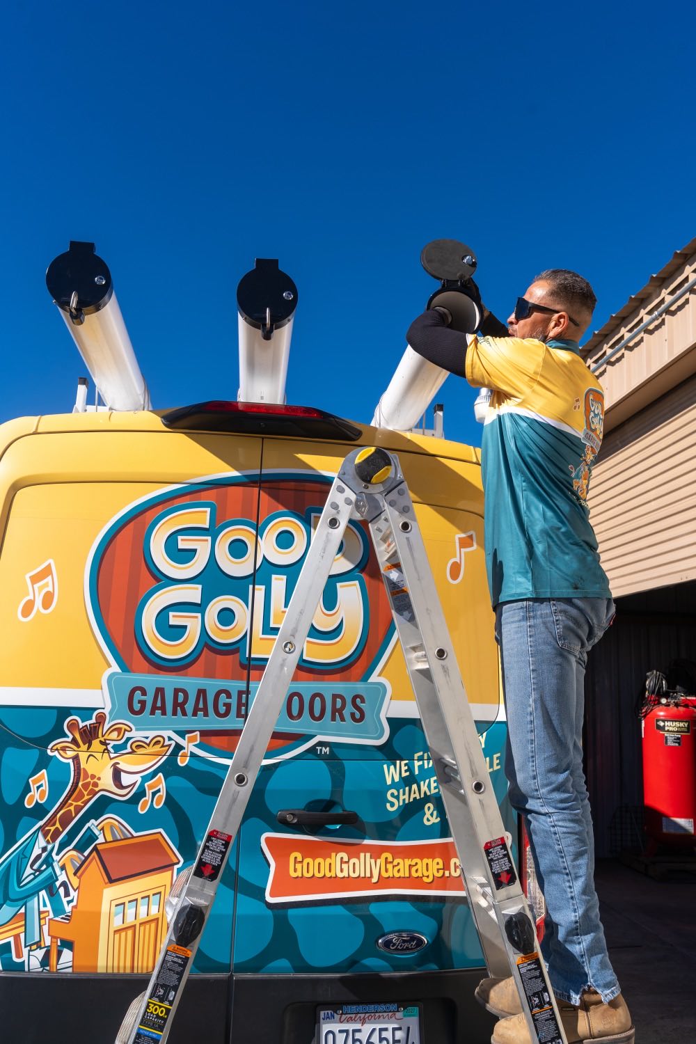 A Good Golly technician on a ladder loading garage door track tubes onto the roof rack of the Victorville van