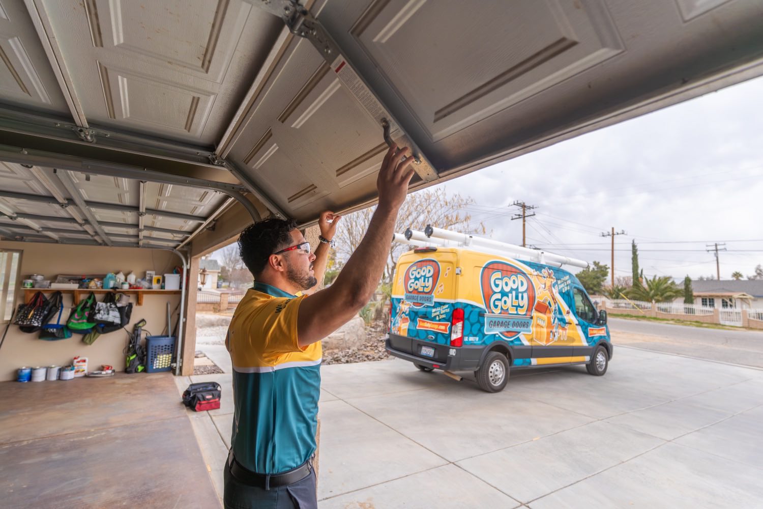 A Good Golly technician checking the top section of an open garage door at a Victorville home, the branded van on the driveway behind him