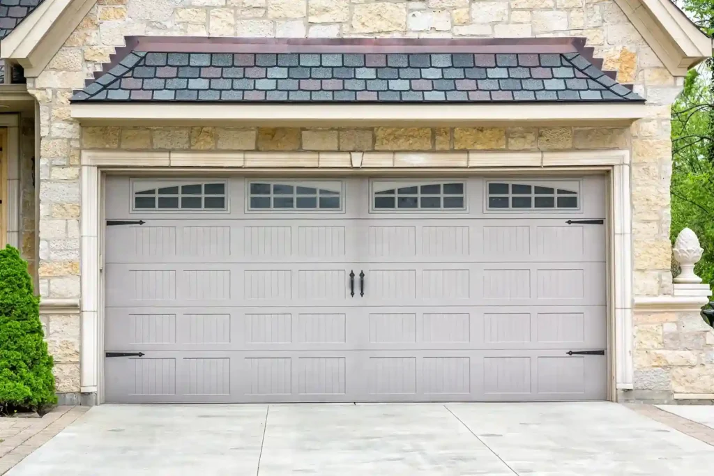 A light gray paneled garage door with decorative black hardware and arched windows, set in a stone and brick facade with a tiered roofline.