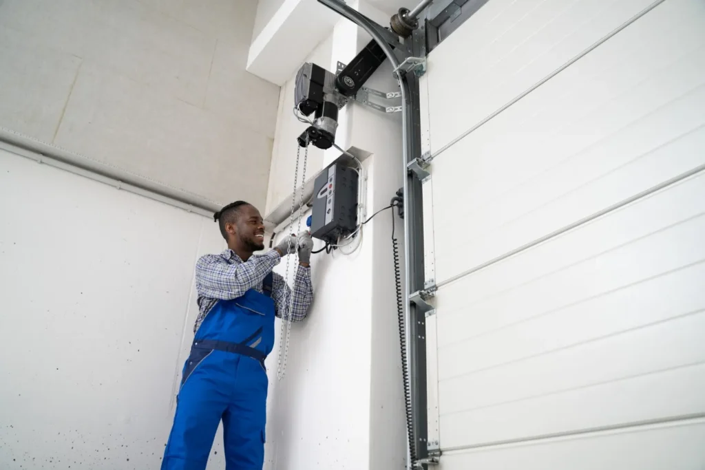 Technician in blue overalls adjusting overhead garage door motor unit near ceiling with tools and gloves.
