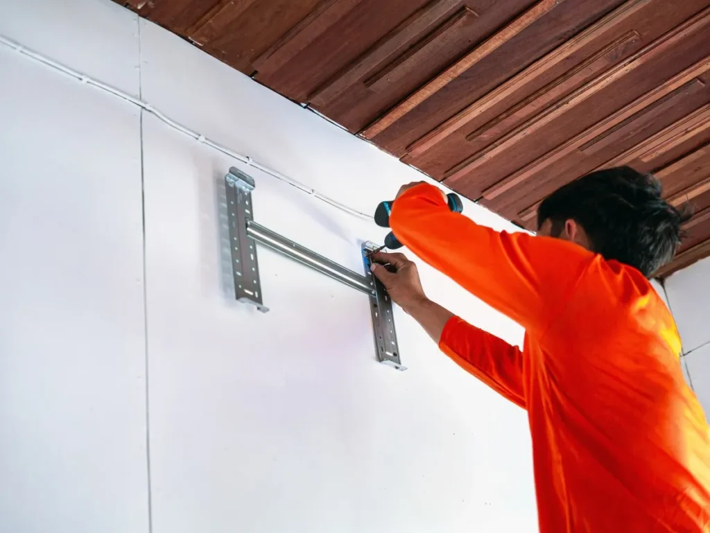 Person in orange shirt using power drill to install metal wall mount bracket on white wall.