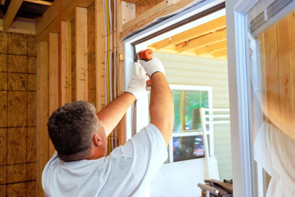 Person in light shirt and gloves using power drill to secure door frame in building under construction.