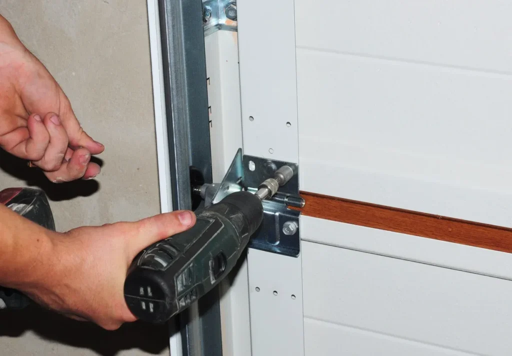 Person using power drill on metal bracket of white sectional garage door. Hands positioned for precision.
