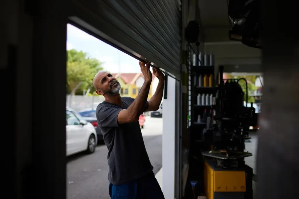 A man manually lifts a rolling metal garage door while looking upward from the outside