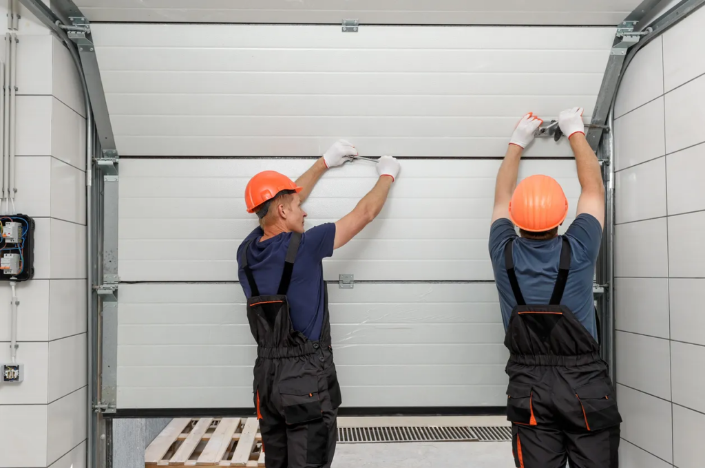 Two technicians wearing orange hard hats and dark overalls work together to install or repair a large white sectional garage door, using hand tools to secure hardware on the upper panels.